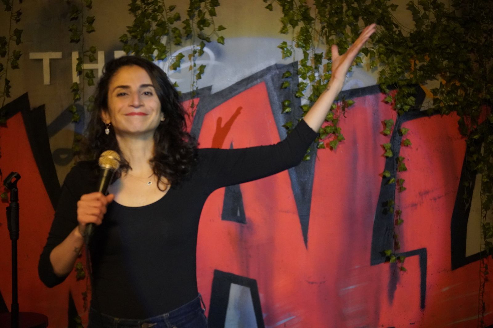 Carmen Chraim on stage in a comedy club, holding a microphone and raising her arm in front of a red graffiti wall