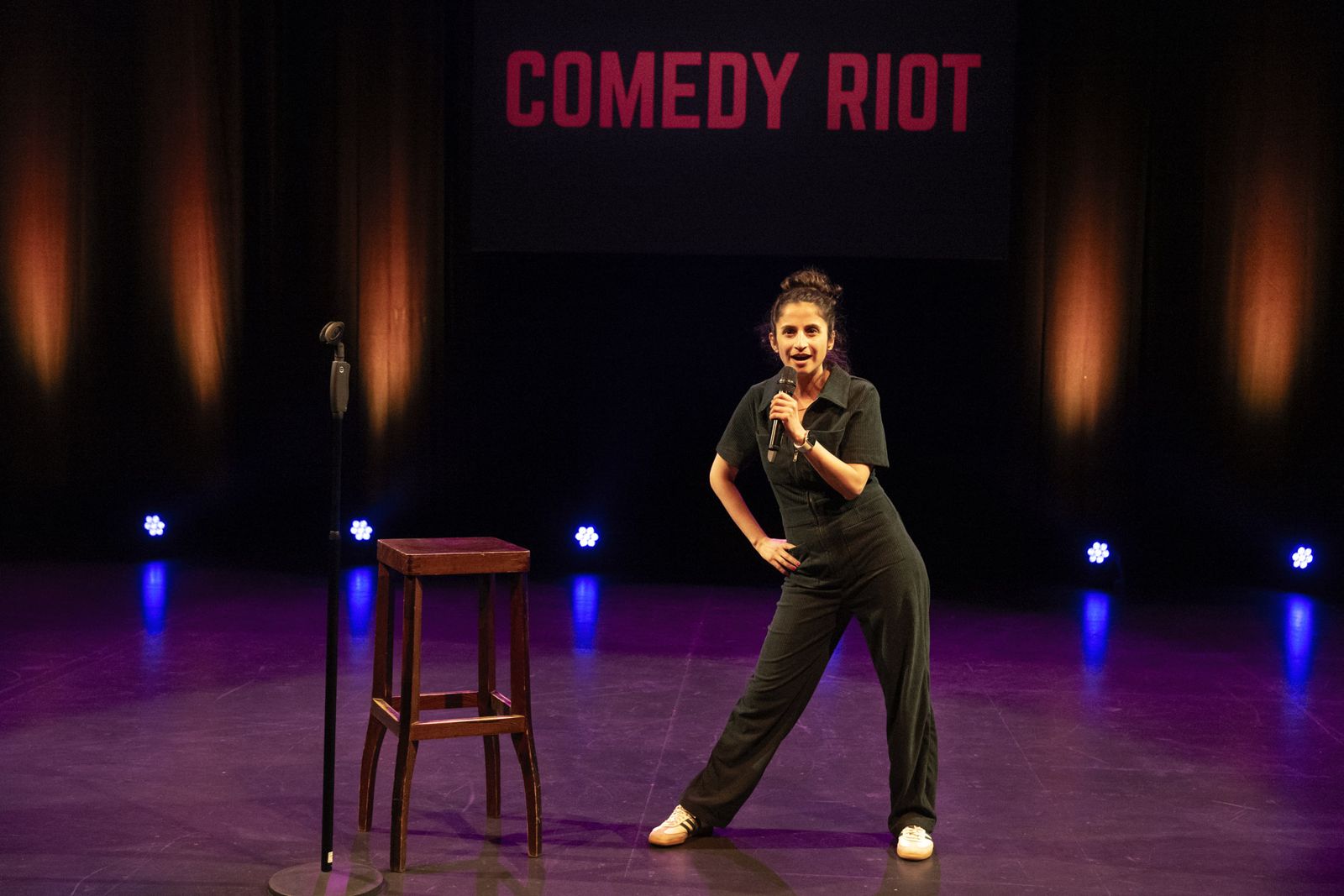 Carmen Chraim performing stand-up on stage with a microphone, next to a wooden stool